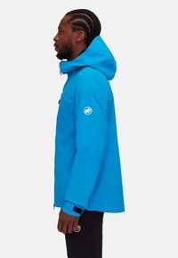 Helle blaue wasserdichte Jacke mit Kapuze, ausgestattet mit einem Frontreißverschluss, kontrastierenden Reißverschlussziehern und einem Logo auf dem linken Ärmel.