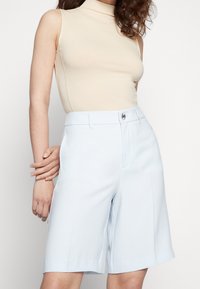 Lichtblauwe op maat gemaakte shorts met een ontspannen pasvorm, voorzien van een tailleband en een sluiting met een knoop aan de voorkant, gecombineerd met een beige mouwloos topje.