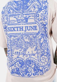 Camiseta beige con un gran diseño gráfico azul, que representa un paisaje urbano con bordes ornamentados y el texto "SIXTH JUNE" destacado.