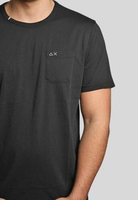T-shirt di cotone nero con collo tondo, maniche corte e taschino sul lato sinistro del petto con un piccolo logo bianco. Texture morbida.