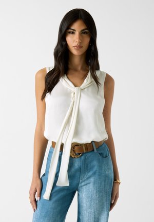 Femme aux longs cheveux bruns porte un chemisier blanc sans manches noué à l'avant, un jean bleu taille haute, une ceinture marron et un bracelet en or.