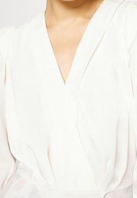 Blusa blanca de manga larga con escote en V profundo, de textura suave y sutil brillo. Las mangas son ligeramente abullonadas, añadiendo dimensión al diseño.