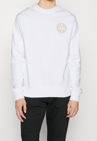 Felpa bianca in cotone, con scollo rotondo, polsini a costine e una toppa circolare con logo beige sul lato sinistro del petto.