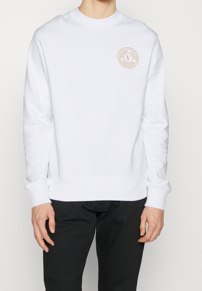 Felpa bianca in cotone, con scollo rotondo, polsini a costine e una toppa circolare con logo beige sul lato sinistro del petto.