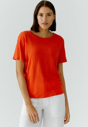 Vrouw met halflang donker haar draagt een feloranje top met korte mouwen en een witte broek, staand tegen een effen lichte achtergrond.