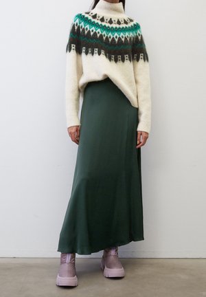 Kremowy sweter z dzianiny z wzorami w kolorach zielonkawym, czarnym i ciemnozielonym, zestawiony z długą ciemnozieloną spódnicą i różowymi botkami za kostkę.