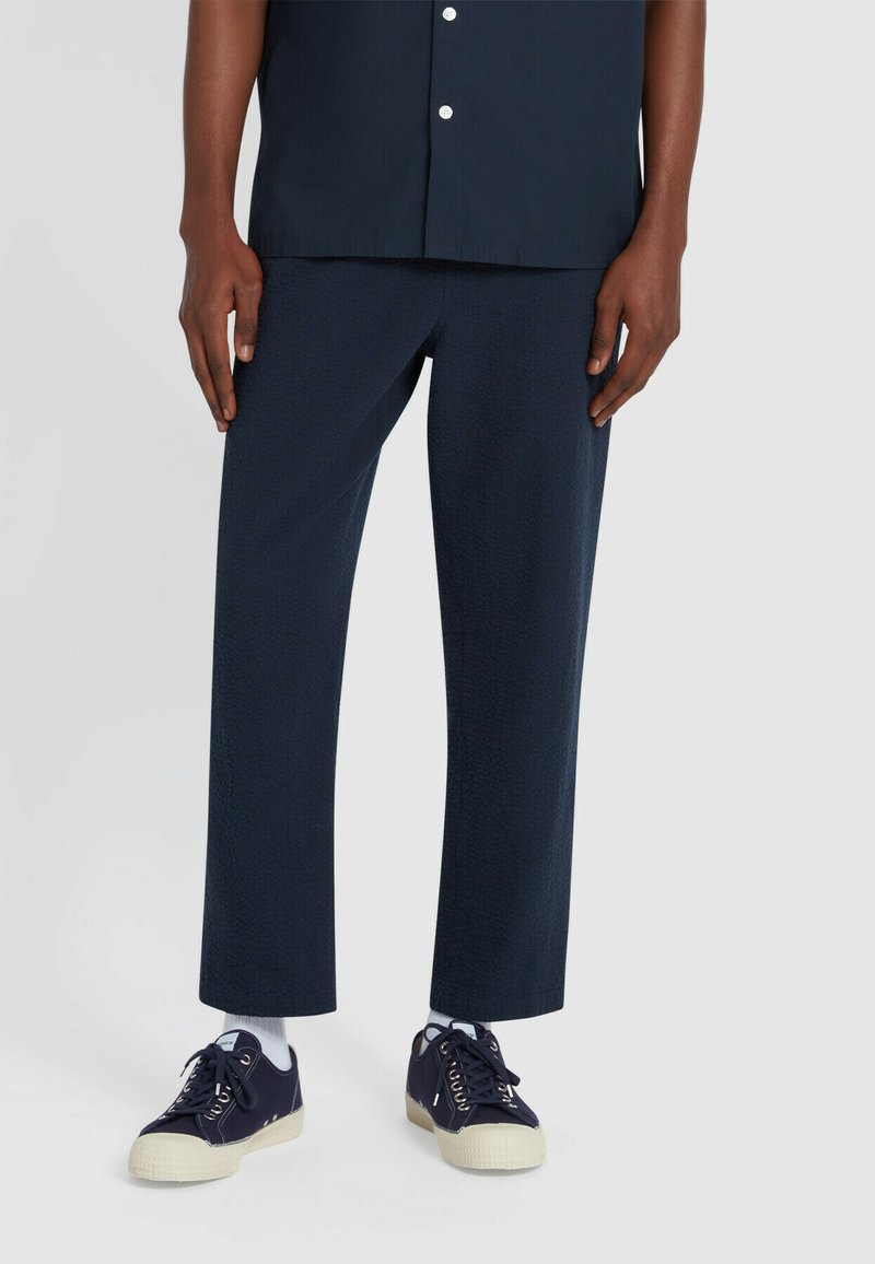 Navy gekleurde textuurbroeken met een recht model, gecombineerd met casual navy sneakers met een crèmekleurige zool. Een overhemd met knopen maakt de outfit compleet.