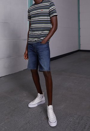 Homme portant des baskets blanches montantes, un short en denim et un t-shirt à manches courtes rayé, debout à l'intérieur contre un mur gris clair.