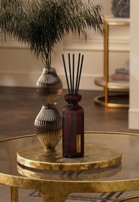 Rød glassdiffuser med svarte stenger ved siden av en ribbet brun vase på et rundt bord i gull. Bakgrunnen inkluderer en uskarp hylle og en plante.