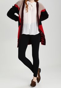 Kvinna klädd i en vit plisserad blus med en slinga, svarta leggings, en lång flerfärgad cardigan och leopardmönstrade tofflor, stående med korsade ben.