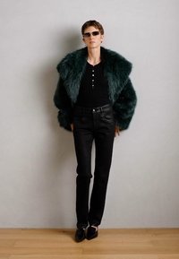 Grön fuskpälsjacka, svart figurnära topp med knappar, svarta jeans och låga skor. Modellen står mot en neutral vägg på ett trägolv.