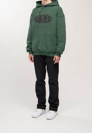 Sudadera verde con "PERSEGUIMIENDO SUEÑOS" impresa en letras negras en negrita. Combinada con jeans negros y zapatillas blancas. El tejido parece suave y casual.
