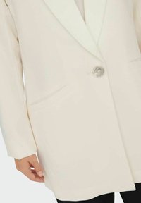 Blazer de couleur crème avec une fermeture à un bouton, deux poches avant et une texture de tissu lisse avec un éclat subtil.