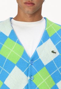 Hombre vistiendo un cárdigan abotonado de rombos en azul, verde y blanco sobre una camiseta blanca de cuello redondo, con un pequeño logo de cocodrilo en el pecho.