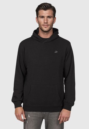 Młody mężczyzna z krótkimi włosami, ubrany w czarną bluzę z kapturem z małym białym logo i podartymi szarymi dżinsami, stojący na tle jednolitego szarego tła.