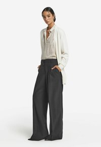 Krämfärgad kavaj över en ljus mönstrad blus och högmidjade byxor i kolgrått med vida ben. Släta texturer och spetsiga klackar.