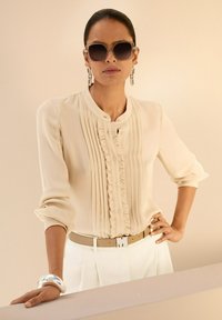 Beige Bluse mit Falten und Rüschen, Knopfverschluss, getragen mit weißen Hosen und einem Gürtel mit einem metallischen Buchstaben. Silbernes Armband als Akzent.