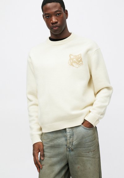 Fiatal férfi fehér kötött pulóverben, bal mellkasán bézs róka logóval és kopott kék farmerban, pózolva, egyik kezét zsebébe téve.