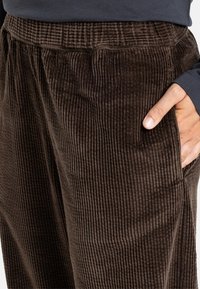 Bruine corduroy broek met verticale ribbelstructuur, elastische tailleband, zijzakken en een ontspannen pasvorm. Close-up zicht.