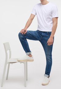 Bílý tričko s krátkým rukávem, modré džíny s ohrnutými manžetami a bílé tenisky s béžovými podrážkami, pózují na bílé židli.