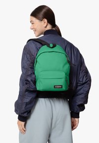 Grön Eastpak-ryggsäck med en texturerad yta, framficka, dragkedja och justerbara remmar, bärs över en marinblå bomberjacka och grå byxor.