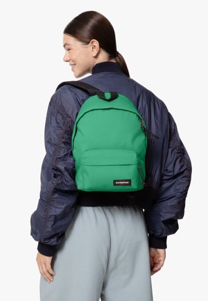 Zaino Eastpak verde con finitura testurizzata, tasca frontale, zip e spallacci regolabili, indossato su una giacca bomber blu navy e pantaloni grigi.