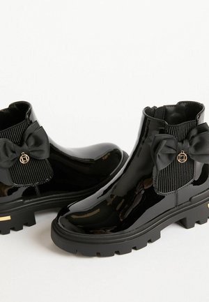 Schwarze Lackknöchelstiefel mit einem gerippten Stoffeinsatz, ausgestattet mit einer großen schwarzen Schleife und goldfarbener Logo-Akzent, strukturierte Gummisohle.