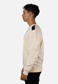 Sudadera beige con detalle negro en el hombro, cuello redondo y mangas largas. Confeccionada en tela suave con un corte clásico.