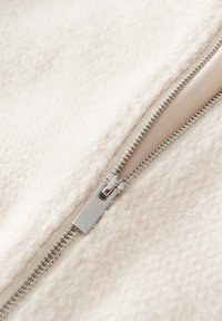 Tela blanca y texturizada con un detalle de cremallera. La cremallera es de metal con un acabado plateado, situada en el borde de la tela, parcialmente abierta.