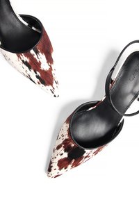 Des talons aiguilles avec des brides en cuir noir et un motif en cuir de vache présentant des taches blanches et brunes. Texture lisse, silhouette élégante.