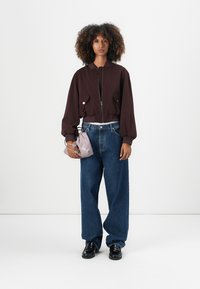 Burgunderfarget cropped jakke med store lommer, sammen med vide blå jeans. Modellen holder en lys lilla veske og har på seg svarte sko.