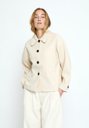 Žena so svetlo hnedými vlasmi, oblečená v krémovom zapínacom saku a voľných bielych nohaviciach, stojaca s rukami v kapsách na bielom pozadí.