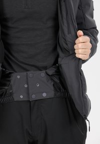 Schwarze isolierte Jacke mit Reißverschluss, Druckknöpfen und elastischem Taillendetail. Glatte Textur mit einem figurnahen Design. Sichtbare Innenauskleidung.