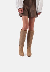Stivali marroni al ginocchio con motivo laserato, parte superiore testurizzata, tacco solido e cinturino alla caviglia. Abbinati a pantaloni shorts marroni e una blusa bianca.