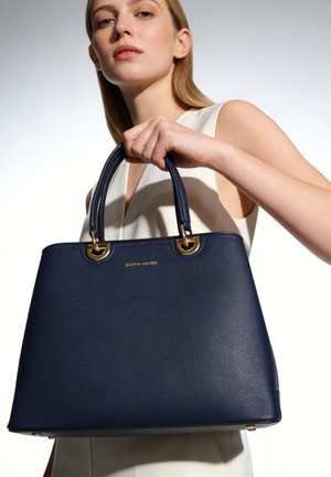 Femme en haut blanc sans manches tenant un sac à main en cuir bleu marine Pierre Cardin avec une quincaillerie dorée, sur un fond clair uni.