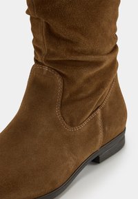 Botte en daim marron hauteur genou avec tige froncée, détails de couture subtils et un petit talon noir. Texture lisse et finition douce.