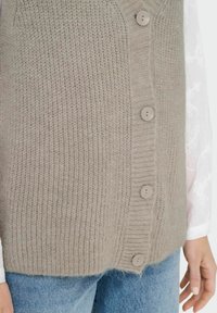 Gilet senza maniche lavorato a maglia beige con una texture a coste, dotato di cinque bottoni grigio chiaro sul davanti. Indossato sopra una camicia bianca a maniche lunghe.