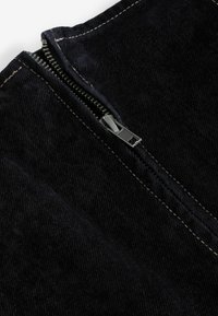 Black corduroy fabric featuring a metal zipper with silver-toned hardware, detailed with contrasting beige stitching along the edges.