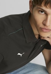 Czarna koszulka polo z białym logo, z turkusowymi przeszyciami na kołnierzu. Nosi ją model z krótkimi włosami.