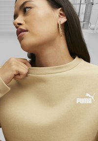 Felpa beige con collo a costine, caratterizzata da un logo PUMA in bianco. Il modello sistema il colletto con una mano. Orecchini a cerchio dorati visibili.