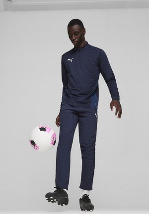 Navy Sportpullover mit Reißverschluss und Seitenpaneelen, kombiniert mit passenden navyfarbenen Hosen. Schwarze Fußballschuhe. Hält einen weißen und rosa Fußball.