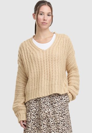 Moteris dėvi smėlio spalvos storą megztinį ant balto viršaus ir leopardo rašto sijoną, stovinti prie paprasto fono.