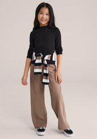 Hosszú ujjú fekete felső széles bézs nadrággal párosítva, a derékon csíkos fekete, fehér és barna megkötős sállal díszítve, fekete tornacipővel viselve.