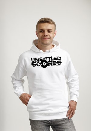 Jonge man draagt een witte hoodie met zwarte tekst "Unsettled Scores Records" en een vinylplaatafbeelding, handen in de zakken, kijkt zijwaarts.