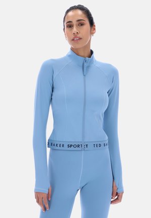 Kvinne iført lys blå treningsjakke med glidelås og lange ermer, og matchende tights med «Ted Baker Sport»-midjebånd, stående foran hvit bakgrunn.
