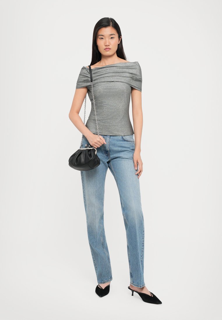 Haut gris à épaule dénudée avec un tissu froncé, associé à un jean droit bleu clair. Le mannequin tient un petit sac à main noir. Escarpins noirs à talons pointus.