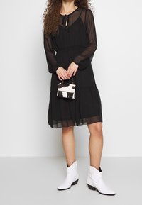 Kvinna i en knälång svart genomskinlig klänning, som håller en liten svart-vit mönstrad handväska och har vita ankelstövlar på sig.