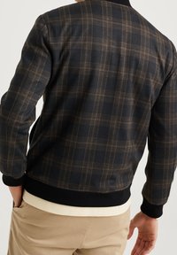 Muž v tmavě kostkované bundě s černými manžetami, oblečený do béžových kalhot, zobrazený z boku s jednou rukou v kapse.