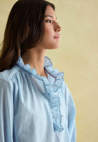 Svetlomodrá, pruhovaná blúzka s volánovým golierom a predným zapínaním. Mäkká tkanina s voľným strihom a jemnou textúrou.