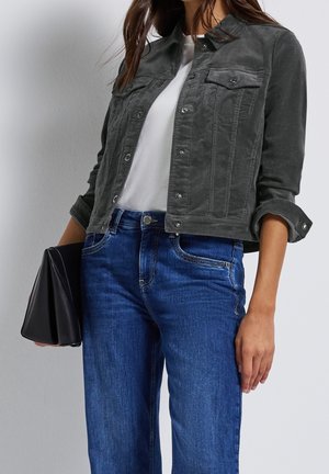 Kvinde iført en hvid skjorte, mørkegrå knappet jakke, blå jeans og holder en sort foldet clutch mod en ensfarvet lys baggrund.